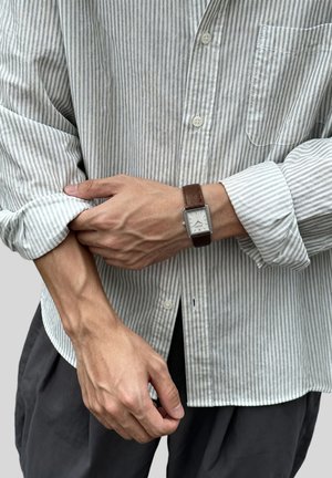 Person trägt ein grau-weiß gestreiftes Hemd und krempelt die Ärmel hoch, mit einer rechteckigen analogen Armbanduhr mit braunem Lederarmband.