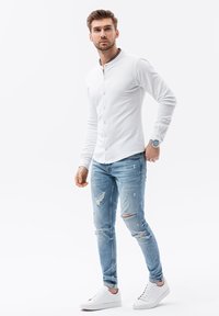Camisa blanca de botones con cuello, combinada con jeans rasgados de color azul claro y zapatillas blancas. El modelo posa contra un fondo plano.