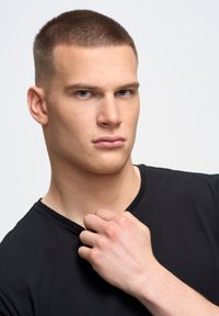 Melns īsām piedurknēm krekls, apaļš kakla izgriezums. Tuvplāns ar vīrieša modeli ar neitrālu izteiksmi, rokas saspiedošas krekla ap kaklu.