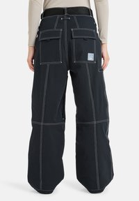 Pantalones de nieve negros con costuras blancas, que presentan una cintura alta, múltiples bolsillos tipo cargo y cremalleras a lo largo de las piernas.