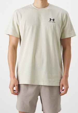 Mann trägt ein schlichtes beiges Under Armour T-Shirt und hellgraue Shorts, steht mit entspannten Armen an den Seiten.