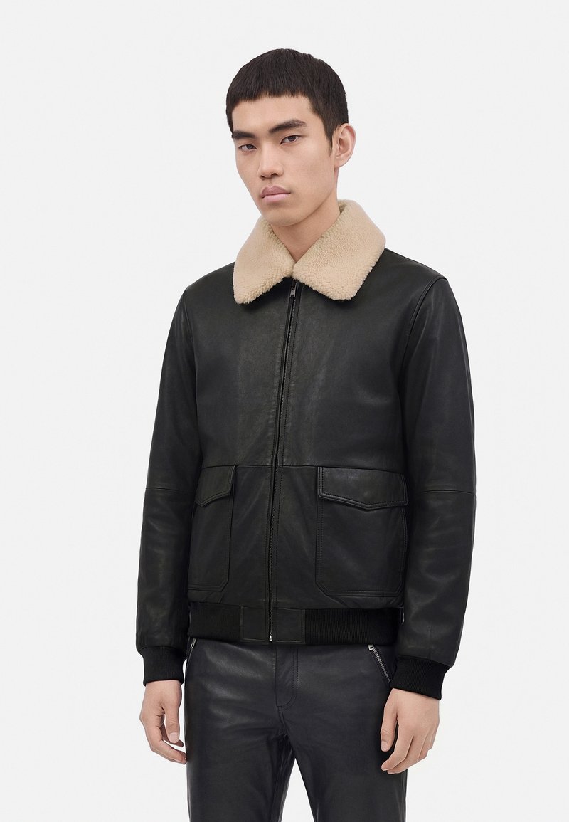 Jeune homme portant une veste en cuir noire avec un col en shearling beige et un pantalon noir, debout devant un fond clair uni.
