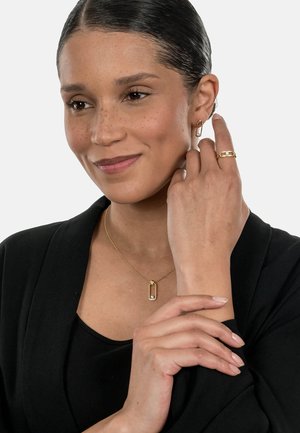 Goldschmuckauslage mit einem Creolenohrring, einem Ring und einer Halskette mit einem rechteckigen Anhänger und Steindetails, vor einem schwarzen Outfit präsentiert.