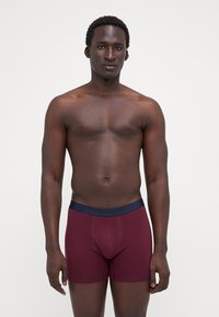 Burgunderfarbene Baumwoll-Boxershorts mit einem marineblauen Bund, die eine glatte Textur und einen enganliegenden Sitz aufweisen, präsentiert an einem Modell vor einem neutralen Hintergrund.