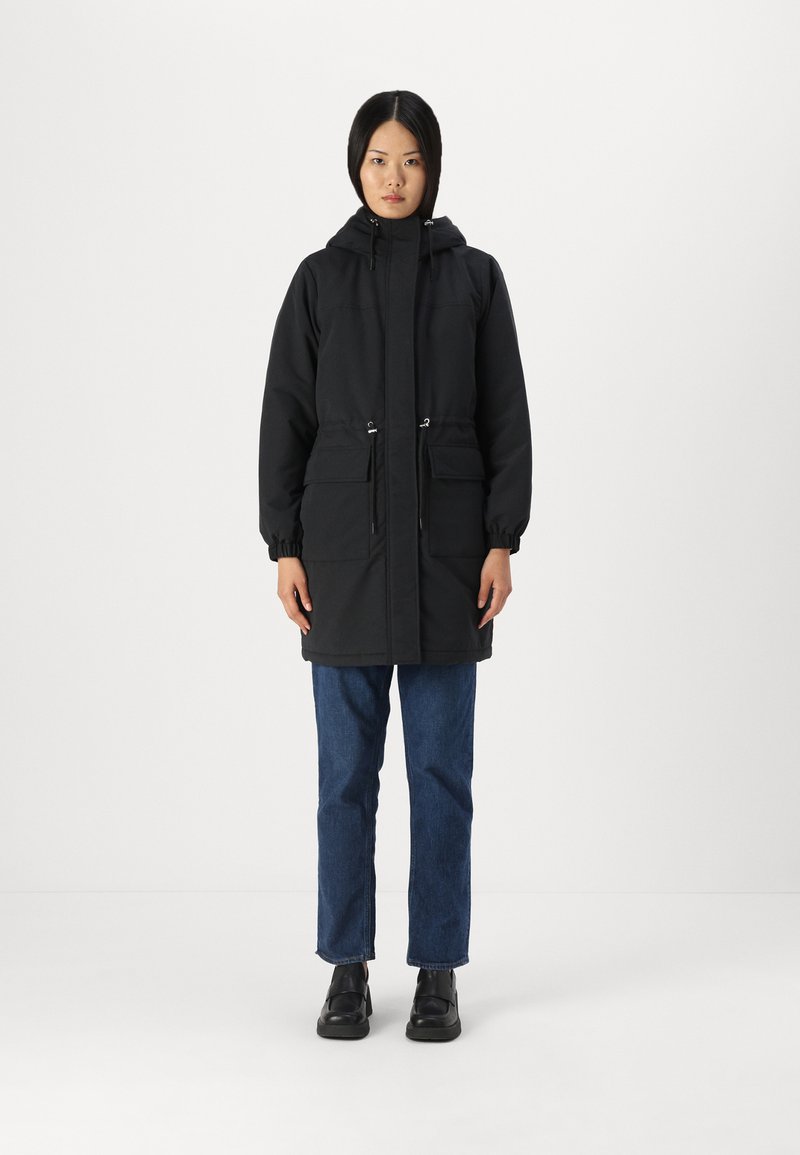 Parka à capuche noire avec deux poches avant, poignets élastiques et cordon de serrage réglable à la taille ; associé à un jean bleu et des chaussures noires.