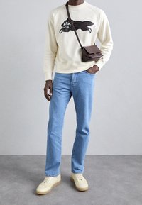 Persona che indossa una felpa crema con un grafiche nero di un coniglio, jeans azzurri chiari, sneakers crema e una piccola borsa a tracolla marrone scuro.