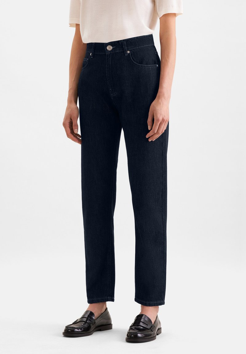Personne portant un jean noir taille haute à jambes droites, un haut blanc à manches courtes et des mocassins en cuir noirs sur un fond blanc uni.
