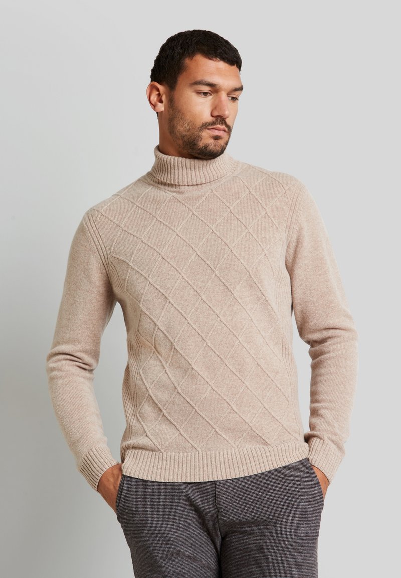 Maglione beige con collo alto, caratterizzato da una trama a diamante e polsini e orlo a coste. Abbinato a pantaloni grigi con texture.
