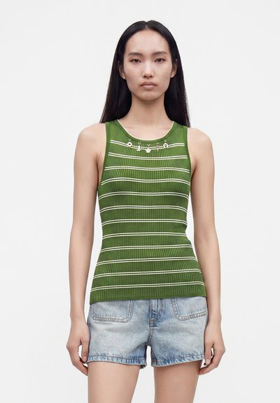Femme aux longs cheveux noirs portant un haut sans manches rayé vert et blanc et un short en jean bleu clair, debout devant un fond uni.