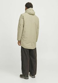 Lys beige vandtæt jakke med hætte, der har en lige pasform og lange ærmer, parret med brede sorte bukser og udendørs fodtøj.