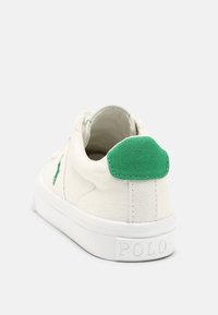 Zapatillas de lona en blanco roto con un acento en tela verde en el talón y una suela de goma blanca texturizada, con un logo en la parte posterior.