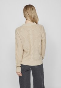 Beige kabelstickad tröja med diamantmönster och texturerad stickning; ribbade mudd och nederkant. Modellen är vänd bort.