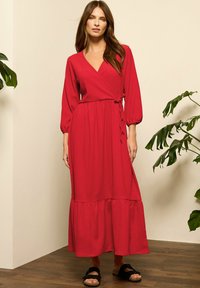Vestido envolvente rojo de manga larga con un dobladillo escalonado, hecho de una tela texturizada. Presenta una cintura ceñida con cinturón y se combina con sandalias planas negras.