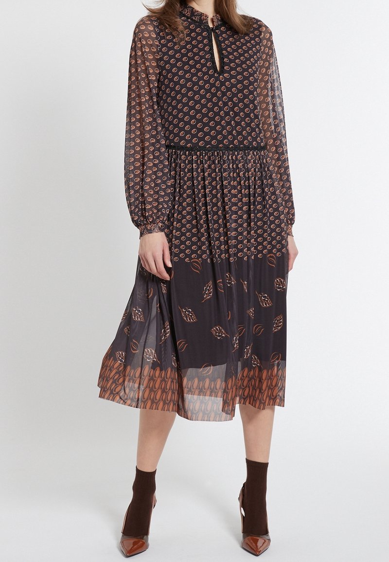 Svart klänning i genomskinligt tyg med långa ärmar, som har en nyckelhålsringning och ett mönster av bruna löv och ovala former över hela ytan. Längd till mitten av vadmalen.