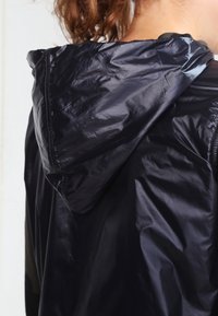 Persona con una chaqueta brillante negra con capucha, vista desde atrás con el cabello castaño suelto parcialmente visible.