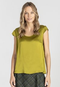 Kobieta o falujących blond włosach ma na sobie błyszcz�ący oliwkowy bezrękawnik i ciemną spódnicę w kratkę, stoi na tle jasnego, jednolitego tła.