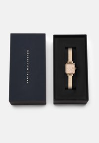 Quadratische goldene Uhr an einem Mesh-Armband, präsentiert in einer mit schwarzem Samt ausgekleideten Box. Die Verpackung hat ein navyblaues Äußeres mit weißen Text.