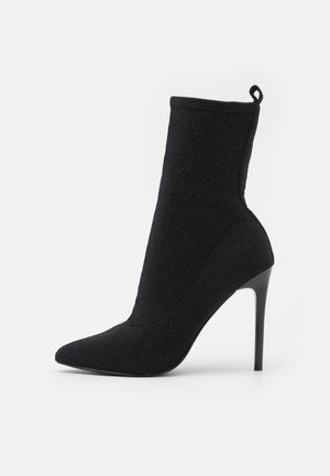 Czarny botek typu sock boot z noskiem w szpic, wysokim obcasem szpilką oraz pętelką z tyłu, wykonany z teksturowanej, elastycznej tkaniny.