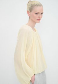 Gelbe durchsichtige Bluse mit runder Ausschnitt und gerafften Ärmeln, kombiniert mit grauen gestreiften Hosen. Weicher Stoff mit fließendem Design.