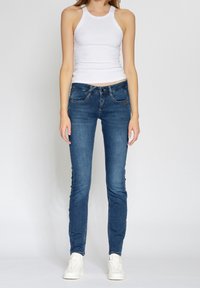 Úzké modré džíny s středně vysokým pasem, pěti kapsami a jemnými švy, kombinované s bílým žebrovaným tílkem a teniskami.