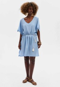 Jasnoniebieska sukienka z dekoltem w serek i luźnymi rękawami, z wiązaniem w talii zakończonym frędzlami. Gładki materiał i długość do połowy uda.