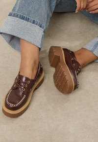 Ruske nahkasaappaat paksulla ruskealla pohjalla, joissa on kultaiset nauhakoristeet ja valkoinen tikkaus. Teksturoitu kumipohja parempaa pitoa varten.