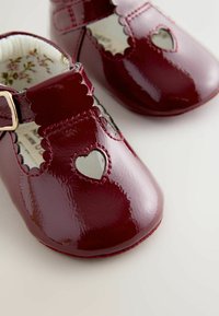 Błyszczące burgundowe niemowlęce buty ze skórki lakierowanej, z falbaniastymi brzegami, wycięciami w kształcie serca oraz bezpiecznym paskiem z złotą klamrą. Kwiatowa podszewka.