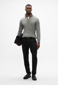 Suéter gris acanalado con cuello de cremallera, pantalones negros, zapatos negros y sosteniendo una chaqueta negra. Diseño simple con tela texturizada.
