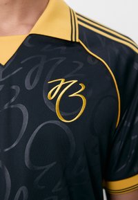 Polo noir avec des accents et un col jaunes. Présente un motif tonal et un logo doré brodé sur le devant. Manches courtes.