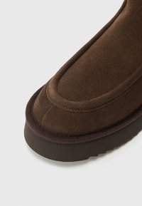 Bruine suède laars met een afgeronde neus, gestikte details langs het bovenwerk en een dikke rubberen zool met een gestructureerde afwerking.