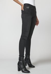 Kvinna i åtsittande svarta jeans med blommotiv och svarta klackboots stående mot en neutral bakgrund.