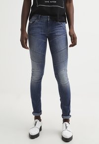 Vaqueros ajustados y desgastados de color azul con textura acanalada en las rodillas, diseño de cinco bolsillos y cintura de altura media. Combinados con una camiseta negra.
