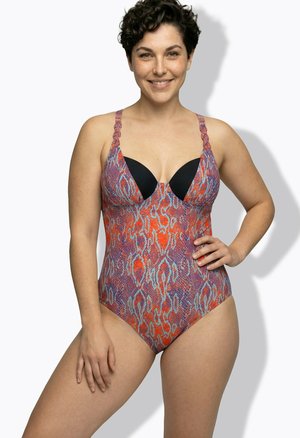 Persona sorridente che indossa un costume intero con motivo serpente arancione e viola e coppe nere con ferretto, mano sui fianchi.