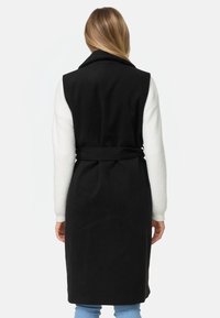 Manteau sans manches noir avec un col haut, ceinture à la taille cintrée et manches longues blanches contrastées. Texture douce, design longueur genou.