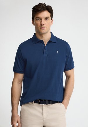 Mężczyzna ubrany w granatową koszulkę polo i beżowe spodnie z paskiem, stoi na tle jednolitego jasnego tła, zwrócony do przodu.