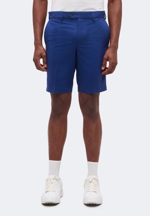 Homme portant un short bleu au genou, des chaussettes blanches et des baskets blanches, debout devant un fond gris clair uni.