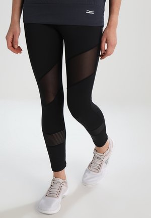 Mujer con mallas negras con paneles de malla y zapatillas deportivas blancas, de pie contra un fondo liso y claro.