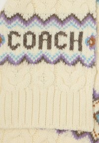 Strikket hat i kremfarvet uld med et zigzag-mønster i lavendel og lyseblå, der tydeligt viser ordet "COACH" i mørkebrun.