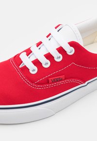 Sneaker in tela rossa con lacci bianchi, linguetta imbottita e etichetta del logo Vans. Presenta una suola in gomma bianca con striscia accentata blu. Superficie testurizzata.