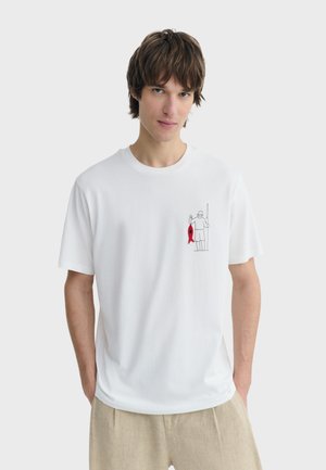 Junger Mann trägt ein weißes T-Shirt mit einer kleinen Fisch-Illustration, hält einen roten Fisch auf der Brust und trägt beige Hosen.