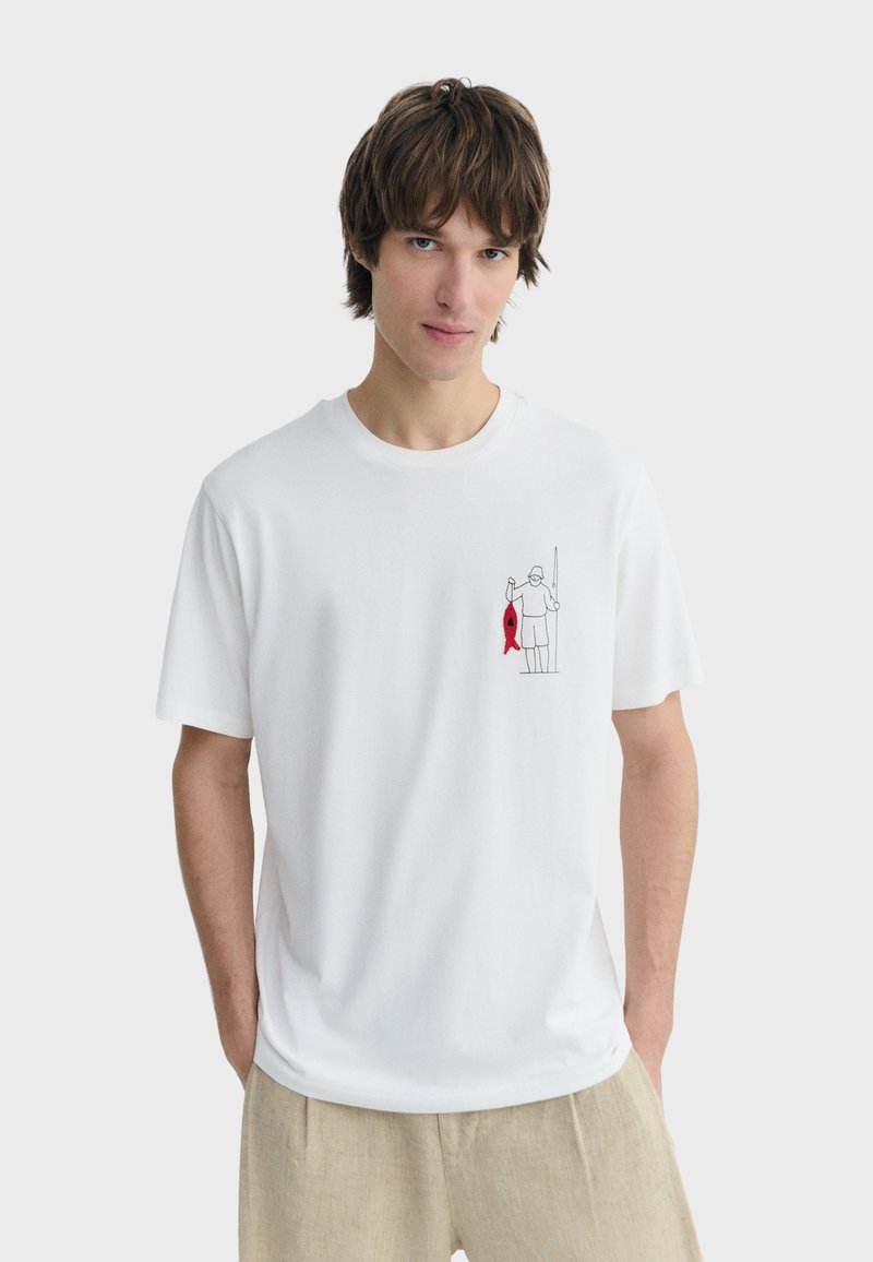 Junger Mann trägt ein weißes T-Shirt mit einer kleinen Fisch-Illustration, hält einen roten Fisch auf der Brust und trägt beige Hosen.