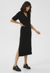 Vestito nero a maniche corte con colletto, spacchi laterali e tasche, realizzato in un tessuto morbido. Abbinato a mocassini neri.