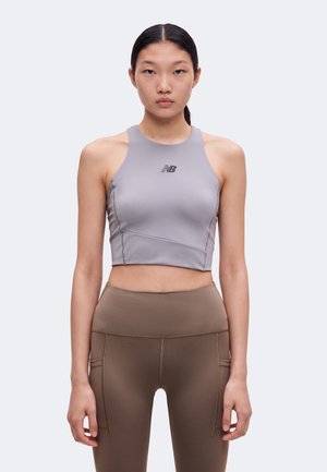 Jeune femme aux cheveux noirs et lisses, portant un crop top gris sans manches et des leggings marron taille haute, faisant face à l'avant sur un fond uni.