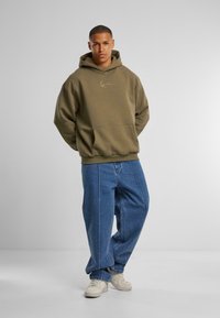 Olijfgroene hoodie met een voorzak, voorzien van een klein geborduurd logo. Gecombineerd met losse blauwe spijkerbroeken en witte sneakers.