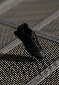 Sapato desportivo preto com o swoosh verde da Nike pairando acima de um chão de grelha metálica em iluminação suave.