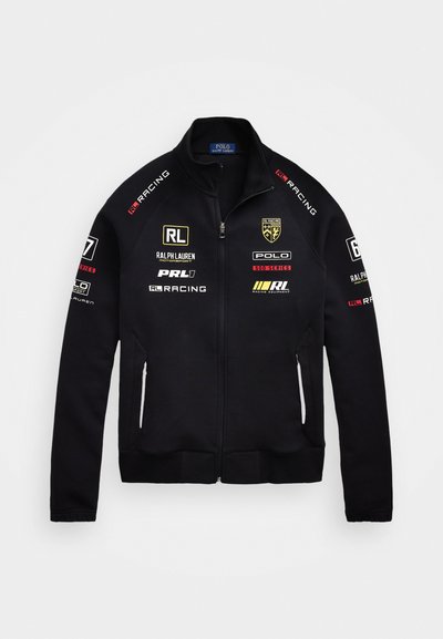 Schwarze Zip-up-Jacke aus weichem Material, versehen mit mehreren Logos in Weiß, Gold und Rot. Sie hat einen hohen Kragen und Seitentaschen.