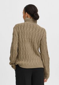 Beige gebreide trui met een hoge kraag, voorzien van een textuurpatroon van verweven vormen. Lange, losse mouwen en een geribbelde zoom.