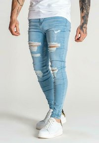 Jeans skinny destrozados de color azul claro, hechos de denim, con múltiples agujeros de desgaste y un diseño ajustado, combinados con zapatillas blancas.