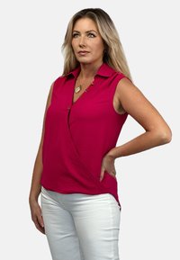 Fuksjowa bluzka bez rękawów z kołnierzykiem, z zapięciami na guziki i krojem w stylu wrap, w połączeniu z białymi spodniami. Tkanina ma teksturowane wykończenie.