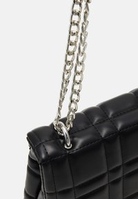 Sac à main en cuir matelassé noir avec un gros plan de sa bandoulière chaîne argentée attachée par un anneau métallique sur un fond clair.